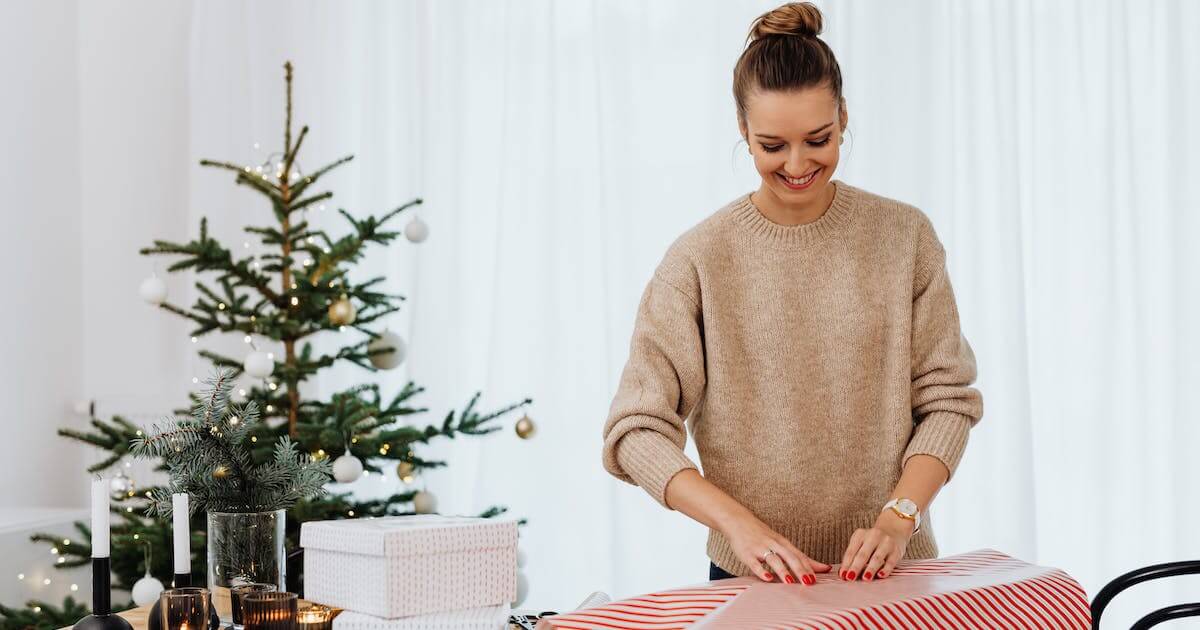 A woman joyfully wraps a gift in front of a beautifully decorated Christmas tree.