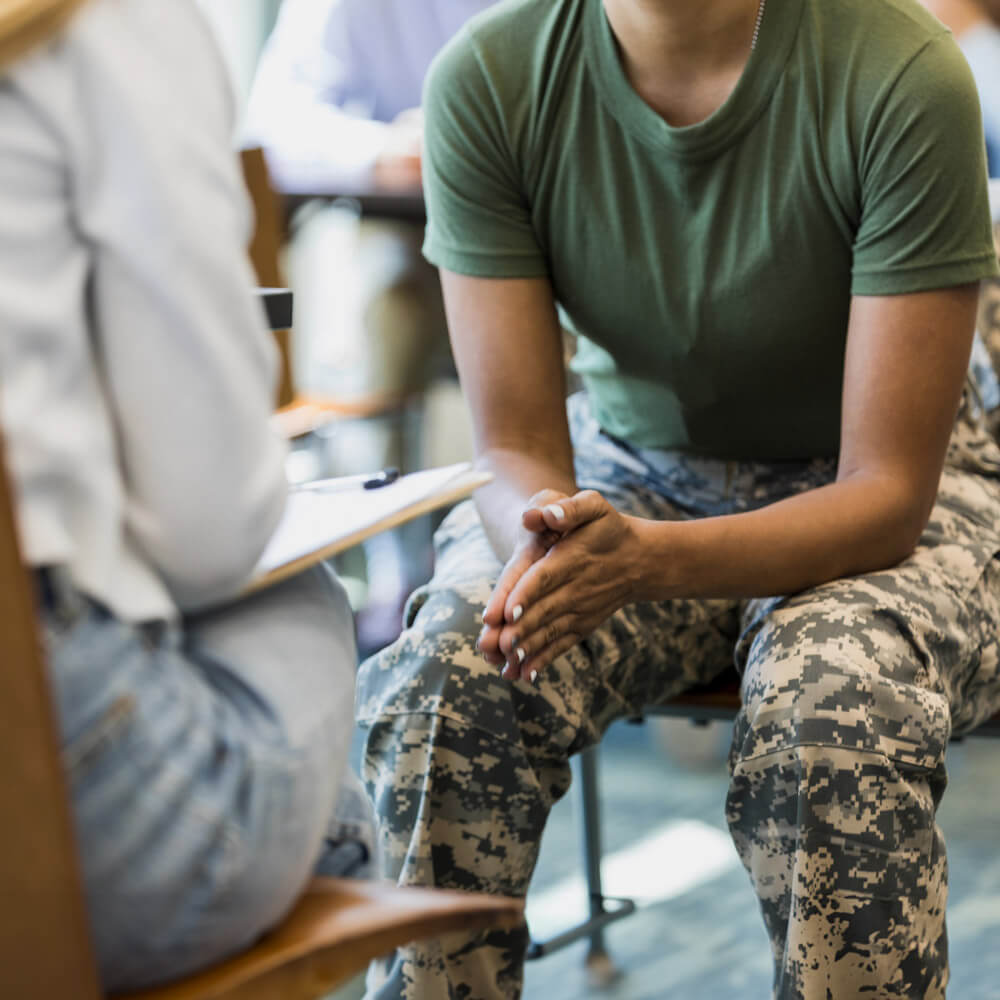 A close up photo of an unrecognizable mid adult female soldier as she puts her hands together and leans forward in her seat. She is talking with an unrecognizable female counselor.