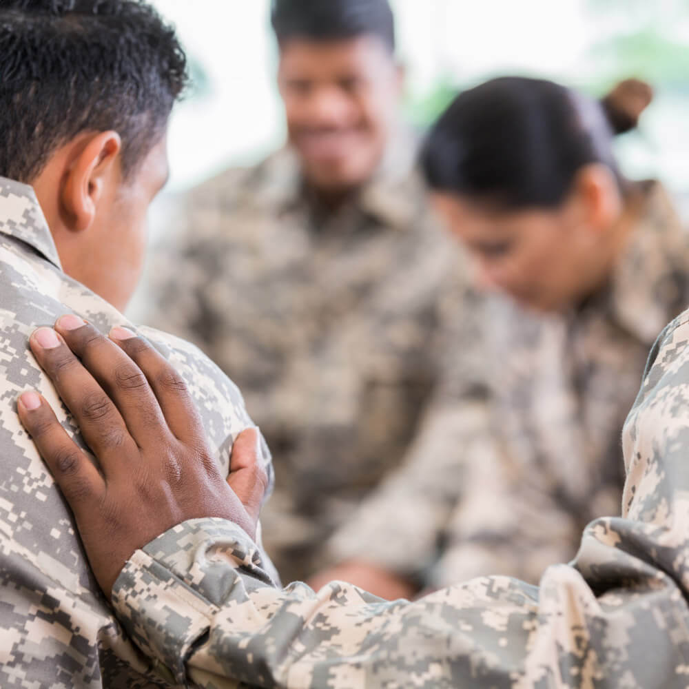 Unrecognizable soldier places his hand on fellow soldier while praying for him during a support group meeting.