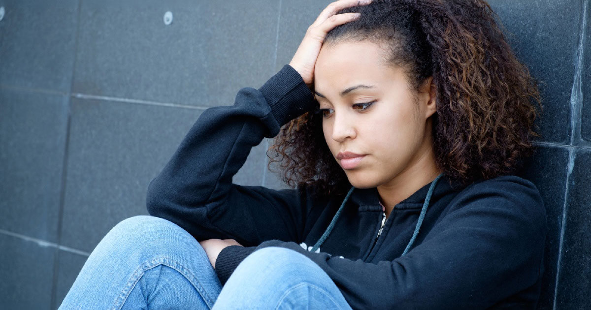 A young woman sits on the ground looking downcast with her hand resting on her head.