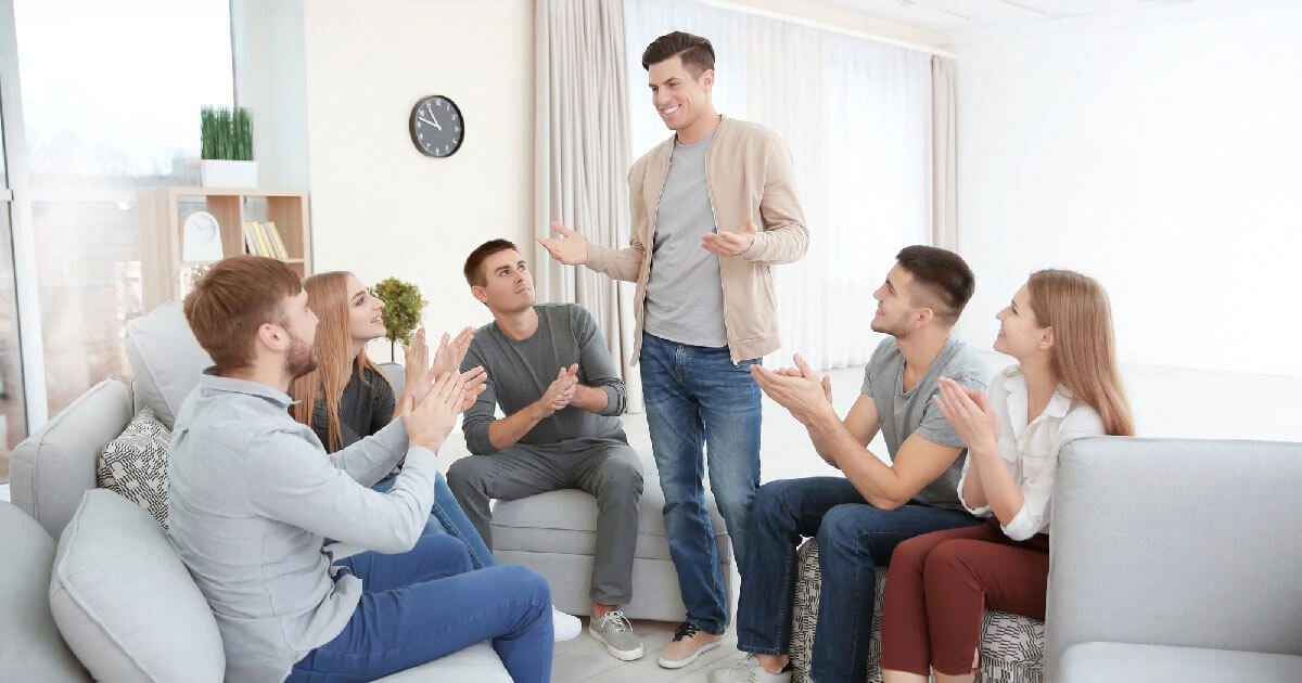 A group of friends sit together clapping and smiling as a man stands speaking to them.